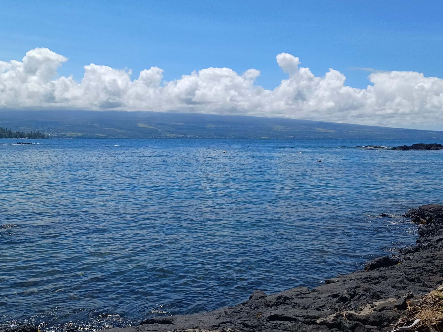 Deep blue bay under a long bank of white clouds, black lava rock along the shore